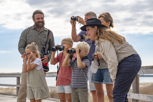 two guides with a family  with three young kids on a boardwalk looking through binoculars and taking a photo.